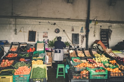 A market stall displaying a variety of fresh vegetables and fruits, including tomatoes, carrots, cucumbers, peppers, onions, and apples. The vendor, wearing an apron, stands behind the stall organizing the produce. The background consists of white tiled walls with hanging decorations, a black clock, and chalkboards.
