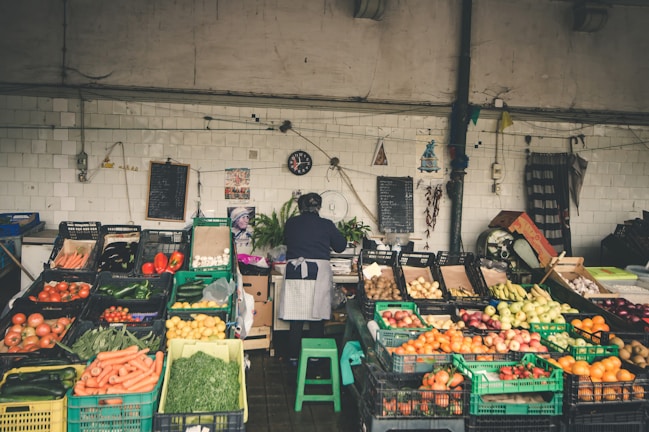 A market stall displaying a variety of fresh vegetables and fruits, including tomatoes, carrots, cucumbers, peppers, onions, and apples. The vendor, wearing an apron, stands behind the stall organizing the produce. The background consists of white tiled walls with hanging decorations, a black clock, and chalkboards.