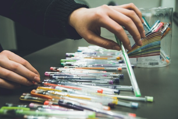 A person is organizing various colorful pens that are spread out on a table. The pens vary in color and are arranged next to a transparent container with a label featuring a striped pattern.