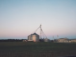 A rural landscape featuring several large metal grain silos with a farm building nearby. In the background, the sky has a soft gradient of colors typical of early evening or dawn. The silos are connected by a network of pipes, and a few utility poles are visible.