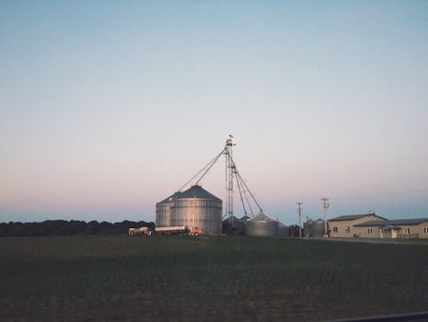 A rural landscape featuring several large metal grain silos with a farm building nearby. In the background, the sky has a soft gradient of colors typical of early evening or dawn. The silos are connected by a network of pipes, and a few utility poles are visible.