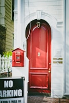 A vibrant red door set in a white brick wall with a decorative arch above it. Adjacent to the door is a vintage red fire alarm box mounted on a white pole. In the foreground, a 'No Parking' sign is partially visible. The scene is well-lit, with shadows indicating a sunny day.