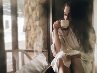 A person with long dark hair is sitting by a window, holding a cup in one hand and a saucer in the other while wearing a robe. The atmosphere appears calm and introspective, with natural light streaming in through the window.