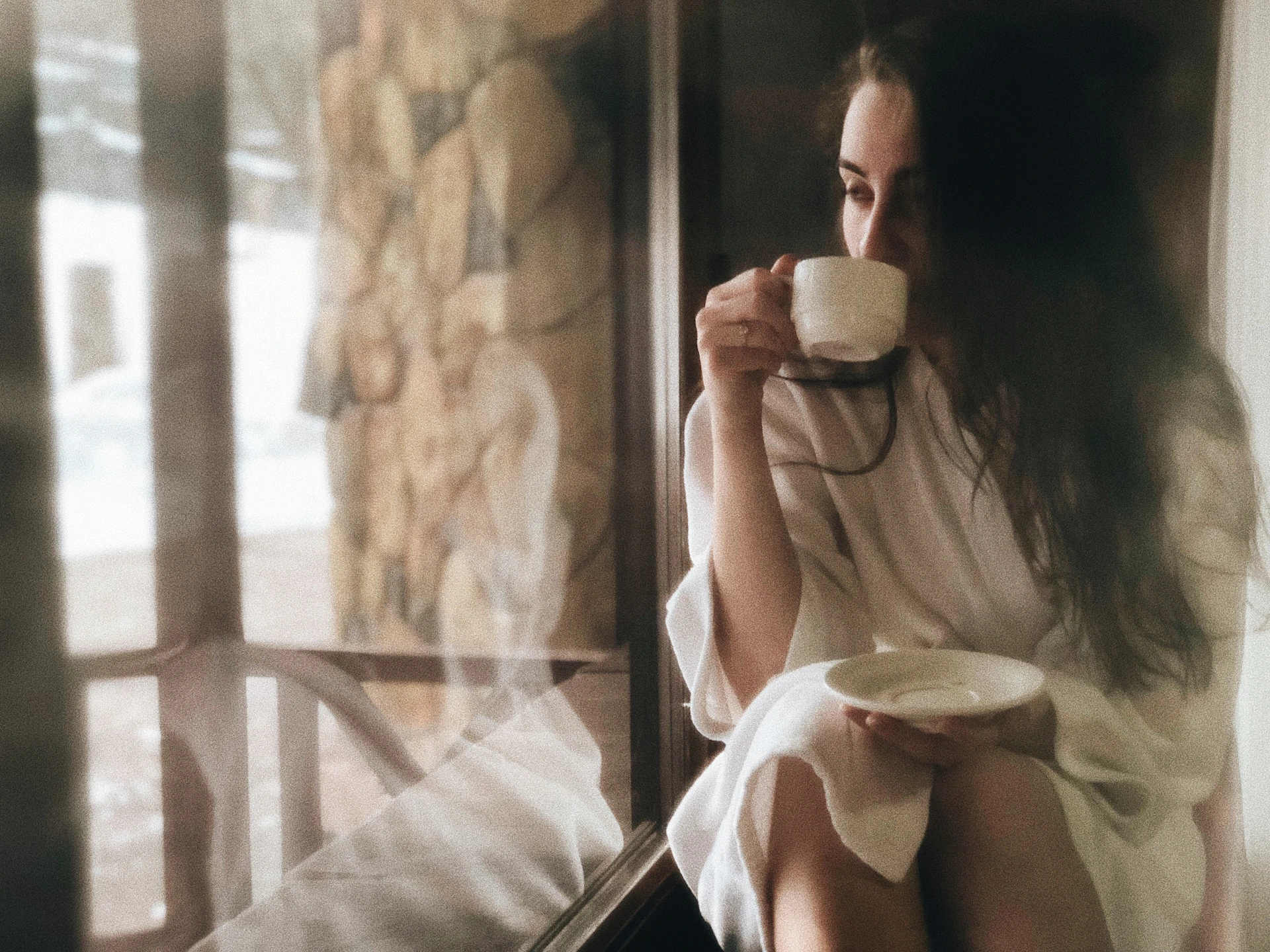 Diana Moon sitting by a window with sheer white curtains, holding a cup of coffee, embodying a relaxed and authentic morning routine.