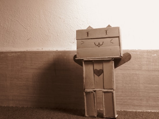 A small cardboard figure resembling a cat stands against a textured wall. It has a simple, happy face with triangular ears on top, stick arms outstretched, and a heart shape on its body. The figure appears homemade, with a sepia tone adding a warm nostalgic feel to the scene.