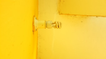 An electric bulb holder is affixed to a bright yellow wall. There is slight shadowing near the connection point, giving depth to the image. The wall texture shows some imperfections and dirt, emphasizing a somewhat aged or utilitarian environment.