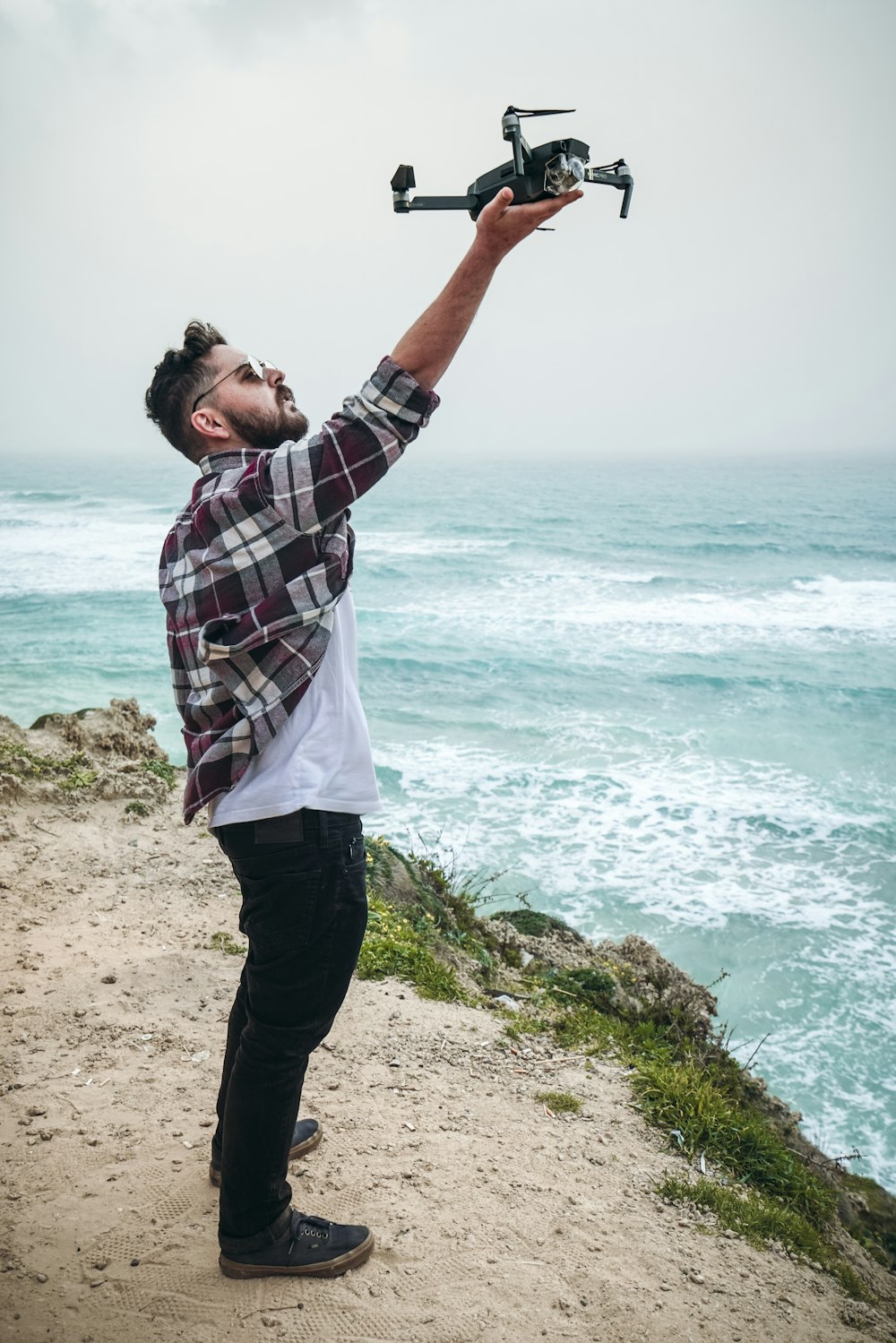 man holding quadcopter while standing on cliff near body of water