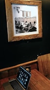 A sepia-toned framed photograph on a dark wall depicting a group of people sitting on benches by a body of water, with a distinctive modern building in the background. Below the photograph, a wooden table holds a laptop displaying a series of thumbnails, with a wooden chair positioned beside it.