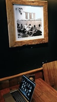 A sepia-toned framed photograph on a dark wall depicting a group of people sitting on benches by a body of water, with a distinctive modern building in the background. Below the photograph, a wooden table holds a laptop displaying a series of thumbnails, with a wooden chair positioned beside it.