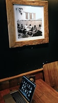 A sepia-toned framed photograph on a dark wall depicting a group of people sitting on benches by a body of water, with a distinctive modern building in the background. Below the photograph, a wooden table holds a laptop displaying a series of thumbnails, with a wooden chair positioned beside it.
