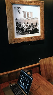 A sepia-toned framed photograph on a dark wall depicting a group of people sitting on benches by a body of water, with a distinctive modern building in the background. Below the photograph, a wooden table holds a laptop displaying a series of thumbnails, with a wooden chair positioned beside it.
