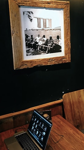 A sepia-toned framed photograph on a dark wall depicting a group of people sitting on benches by a body of water, with a distinctive modern building in the background. Below the photograph, a wooden table holds a laptop displaying a series of thumbnails, with a wooden chair positioned beside it.