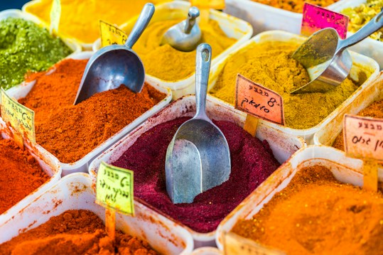 Various colorful spices are displayed in large plastic containers, each filled with vibrant powders like red, yellow, orange, and green. Metal scoops are placed in or near some of the containers. Small handwritten labels with text are stuck into the spices.