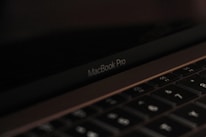 A close-up view of a MacBook Pro laptop screen and keyboard. The focus is on the MacBook Pro label below the screen, with the keyboard keys visible in the foreground. The lighting is low and the atmosphere is subdued, lending a sleek and minimalistic feel.