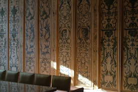 A wall adorned with intricate and ornate decorative panels featuring classical motifs and faces in a vintage style. The panels have detailed floral and leaf patterns with a muted color palette, and sunlight gently illuminates parts of the wall. There is a row of green upholstered chairs lined up against the wall, hinting at a formal or elegant setting.