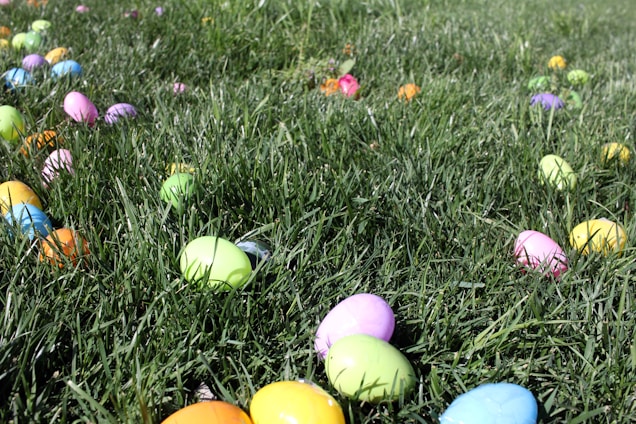 Brightly colored plastic eggs are scattered across a lush green grassy field, suggesting a playful, festive atmosphere likely associated with an Easter egg hunt.
