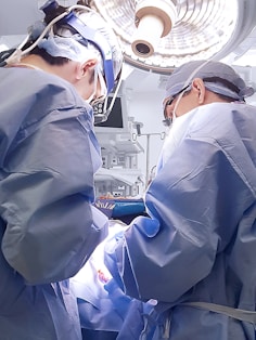 Two surgeons wearing blue surgical gowns and caps perform an operation under bright surgical lights. Medical equipment and monitors surround them in a sterile operating room environment.