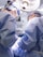 Two surgeons wearing blue surgical gowns and caps perform an operation under bright surgical lights. Medical equipment and monitors surround them in a sterile operating room environment.