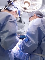 Two surgeons wearing blue surgical gowns and caps perform an operation under bright surgical lights. Medical equipment and monitors surround them in a sterile operating room environment.