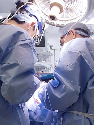Two surgeons wearing blue surgical gowns and caps perform an operation under bright surgical lights. Medical equipment and monitors surround them in a sterile operating room environment.
