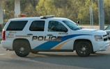 A police SUV with the words 'Honor Pride Dignity' on the side drives along a road. The vehicle is white with blue and gold stripes and has tinted windows. It is in motion near a forested area and a gas station.