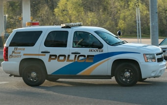 A police SUV with the words 'Honor Pride Dignity' on the side drives along a road. The vehicle is white with blue and gold stripes and has tinted windows. It is in motion near a forested area and a gas station.
