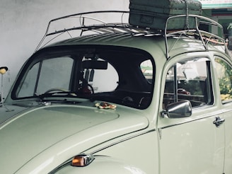 A vintage light green car with a roof rack carrying a weathered suitcase. The car is parked under a soft, natural light, highlighting its classic curves and retro design. The background shows a partially visible interior through the rear and side windows.