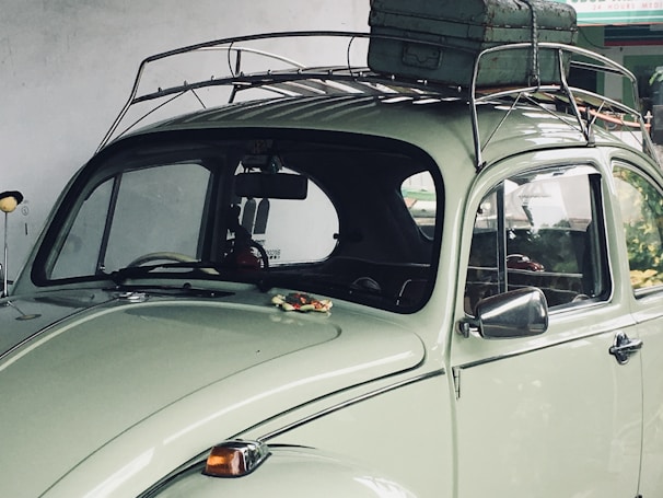 A vintage light green car with a roof rack carrying a weathered suitcase. The car is parked under a soft, natural light, highlighting its classic curves and retro design. The background shows a partially visible interior through the rear and side windows.