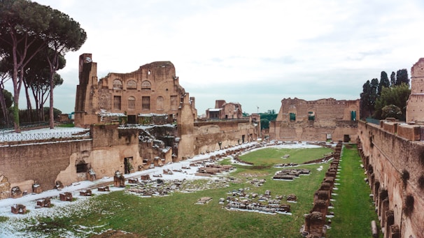 Ancient ruins with remnants of stone structures and archways. The scene is surrounded by tall trees and cloudy skies, with patches of grass and sections of snowy ground.