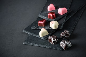 Heart-shaped chocolates in various colors, including pink, red, white, and dark brown, are arranged on black slate plates. The chocolates have a glossy texture, with some featuring speckled patterns.