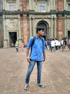 A young person is standing in front of an old, historic building with large stone columns and intricate architectural details. The person is wearing a blue shirt, jeans, and sandals, and has a backpack. Several other people are visible in the background, suggesting a tourist location.