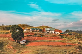 A rural landscape featuring a house with a green roof situated on a hill. Surrounding the house are other small buildings and a few parked vehicles. The area is characterized by red soil, patches of green grass, and a few trees. The sky is bright with scattered clouds, suggesting a clear day.