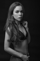 A black-and-white portrait of a young woman with long hair, wearing a black lace bralette and denim jeans. She is posing confidently with one hand raised near her other shoulder, looking directly at the camera.