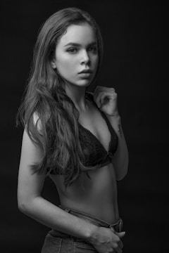 A black-and-white portrait of a young woman with long hair, wearing a black lace bralette and denim jeans. She is posing confidently with one hand raised near her other shoulder, looking directly at the camera.