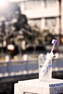 A glass filled with liquid and a toothbrush is placed on a stone surface. The toothbrush has a white handle with purple bristles and a small white flower attached. In the background, there is a blurred view of a building and trees, with bright sunlight creating a soft, glowing effect.