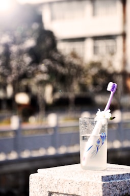 A glass filled with liquid and a toothbrush is placed on a stone surface. The toothbrush has a white handle with purple bristles and a small white flower attached. In the background, there is a blurred view of a building and trees, with bright sunlight creating a soft, glowing effect.