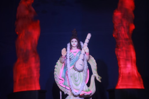 A statue of a figure holding a musical instrument, adorned in colorful garments with a flower garland. The background features abstract red lighting that resembles flames.