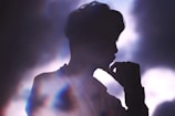 A silhouette of a person is depicted against a backdrop of soft, cloudy light. The person appears contemplative, with their hand resting near their mouth, casting a calm and introspective aura.