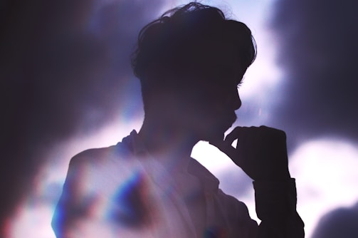 A silhouette of a person is depicted against a backdrop of soft, cloudy light. The person appears contemplative, with their hand resting near their mouth, casting a calm and introspective aura.