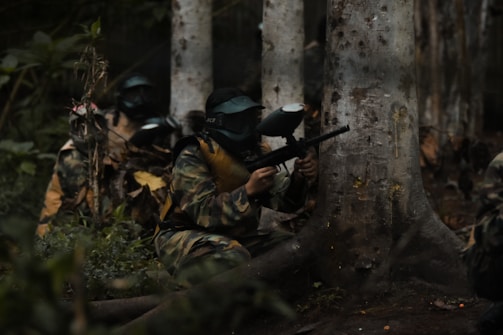 Two people dressed in camouflage and protective masks are crouching behind a tree, holding paintball guns. The setting appears to be a forest with dense foliage, as there's a visible tree trunk and various leaves on the ground.