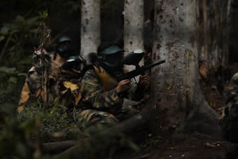 Two people dressed in camouflage and protective masks are crouching behind a tree, holding paintball guns. The setting appears to be a forest with dense foliage, as there's a visible tree trunk and various leaves on the ground.