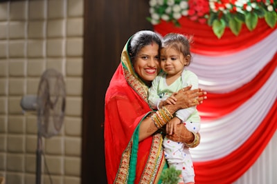 A woman wearing a red saree with gold and green details is joyfully holding a child dressed in white with colorful polka dots. Both have henna designs on their hands, and the background features red and white draped fabric with green and white floral decorations.