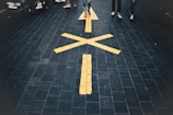 A paved walkway with a large yellow arrow and street markings. People are walking along the path, and the ground appears to be made of dark, textured tiles.