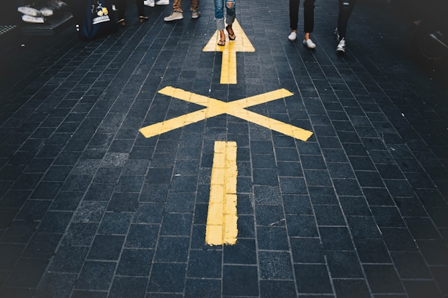 A paved walkway with a large yellow arrow and street markings. People are walking along the path, and the ground appears to be made of dark, textured tiles.