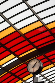 A large clock with simple black hands and no numbers is mounted against a background of a roof made up of red, orange, and white glass panels arranged in rows. The clock has a minimalist design with a modern feel and features red crosses below the center point, situated in a public space that suggests a station or transit area.