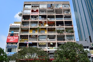 A multi-storey building with numerous small shops, cafes, and businesses, all with colorful signs. The front of the building features various advertisements and shop names, with balconies decorated with plants and some stores displaying items like lanterns. The sky is clear and blue, with some trees visible at the bottom of the image.