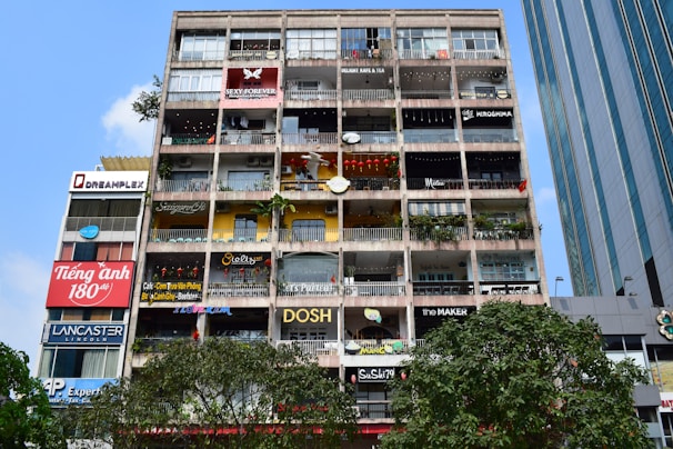 A multi-storey building with numerous small shops, cafes, and businesses, all with colorful signs. The front of the building features various advertisements and shop names, with balconies decorated with plants and some stores displaying items like lanterns. The sky is clear and blue, with some trees visible at the bottom of the image.