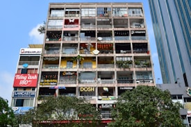 A multi-storey building with numerous small shops, cafes, and businesses, all with colorful signs. The front of the building features various advertisements and shop names, with balconies decorated with plants and some stores displaying items like lanterns. The sky is clear and blue, with some trees visible at the bottom of the image.