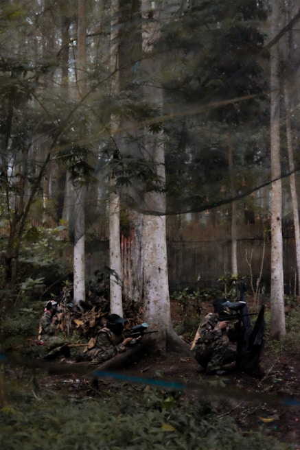 A group of people in camouflage gear and protective masks are engaged in a paintball game in a forested area. The surroundings are dense with tall trees and undergrowth, creating a natural environment suitable for the game.