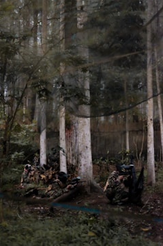A group of people in camouflage gear and protective masks are engaged in a paintball game in a forested area. The surroundings are dense with tall trees and undergrowth, creating a natural environment suitable for the game.